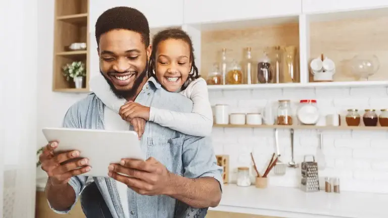 A smiling father looking at a tablet with his daughter hugging him and looking on from behind